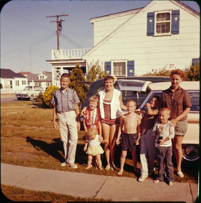2 women pose with 6 children, the year looks to be early-to-mid 1960s. A white house with blue shutters in the background. 