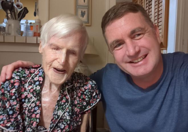 An elderly woman laughing sitting at a dining room table, a young man sits next to her with an arm around her, smiling, looking at the camera while he takes the selfie of the two of them. 